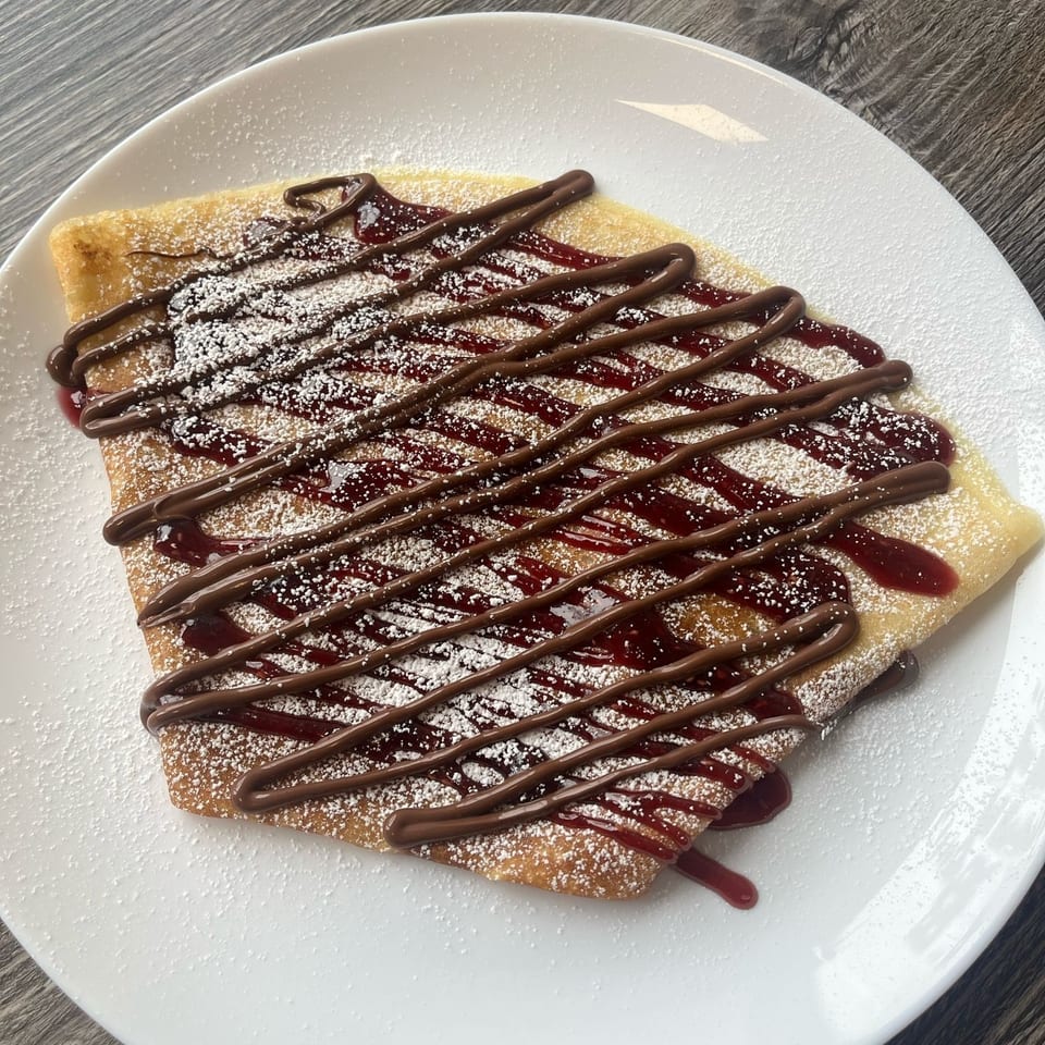 A folded crepe drizzled with chocolate and raspberry sauce, dusted with powdered sugar on a white plate.