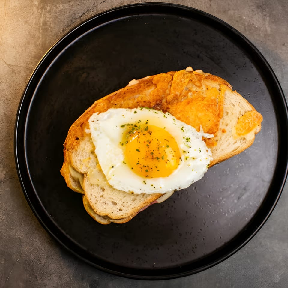 Overhead view of a fried egg with herbs on cheesy toast, served on a black plate.