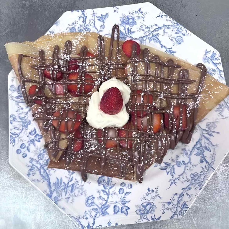 Strawberry and chocolate drizzled crepe with whipped cream and powdered sugar on a patterned plate.